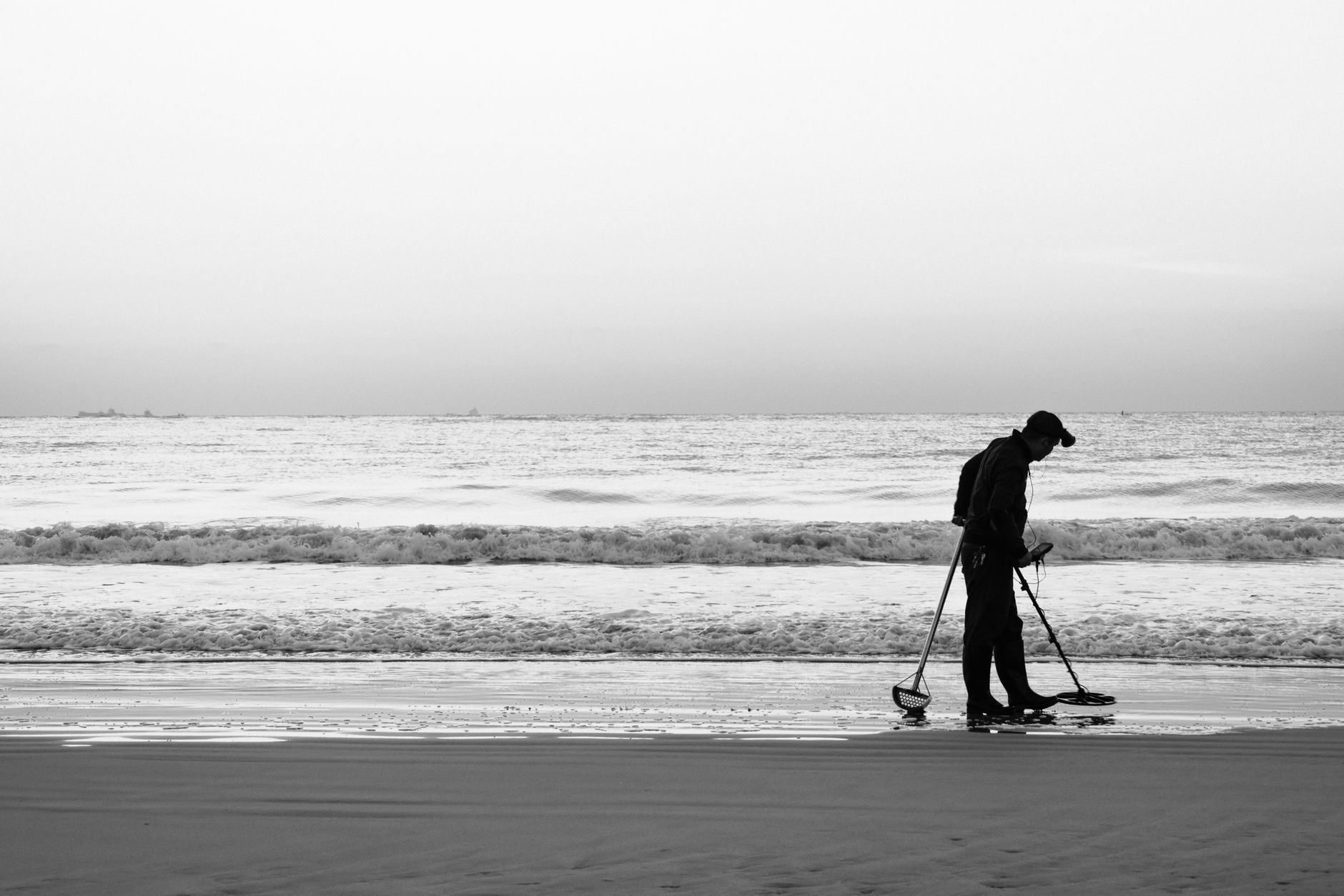 A person walking a quiet beach at dawn holding a metal detector while a partner waits alone near a parked car